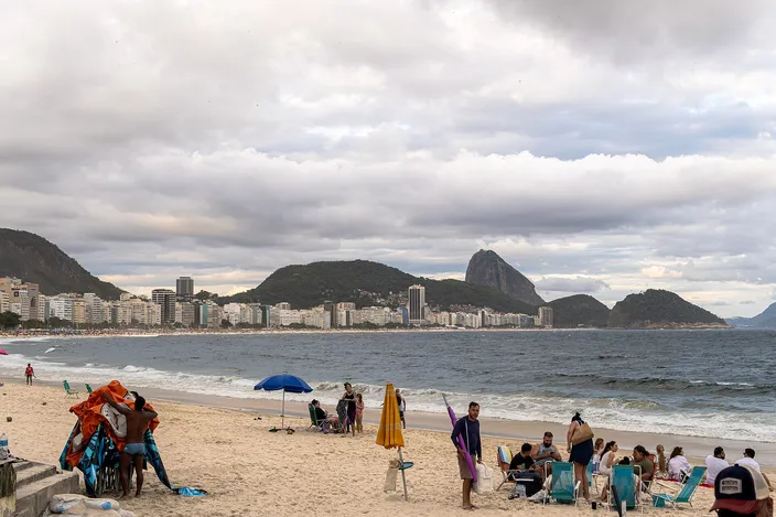 A wide stretch of Copacabana sand seen from Posto 5 on an overcast afternoon — beach umbrellas, deckchairs and a vendor's tent in the foreground, whitewater breaking along the shoreline, and a twin-peaked mountain beyond the water
