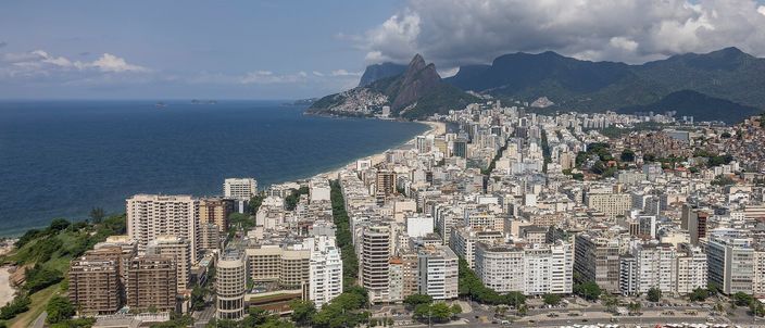 Aerial view along the curving sand of Ipanema beach, dense white apartment blocks lining the shore, with the twin granite summits of Dois Irmãos rising in the distance under a bright blue sky with scattered white clouds