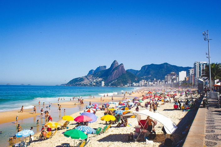 Ground-level view along Ipanema beach on a clear day, packed with striped umbrellas and bathers at the waterline, calm turquoise water lapping pale sand, with the twin peaks of Dois Irmãos visible down the curve of the shore