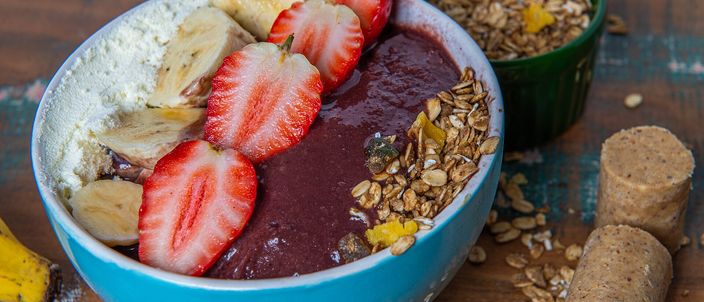A bowl of dark purple açaí topped with sliced banana, strawberry and granola, with more granola alongside