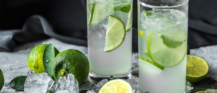 Two glasses of caipirinha with lime slices and ice, surrounded by fresh limes on a dark surface.