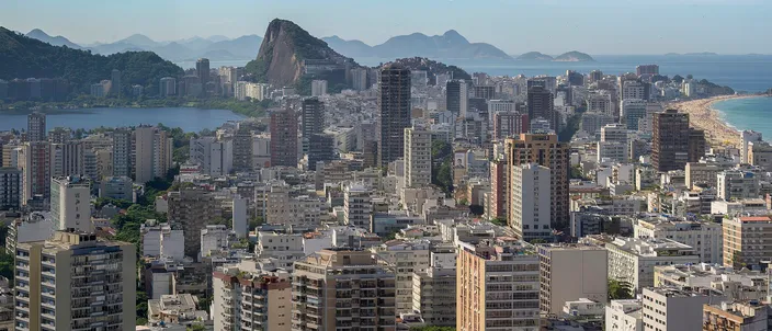 Dense white apartment towers of Rio's Zona Sul filling the frame, with the pointed silhouette of Sugarloaf-style granite peaks and Guanabara Bay behind, and a strip of ocean beach at the right edge under a clear sky