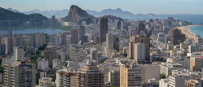 Dense white apartment towers of Rio's Zona Sul filling the frame, with the pointed silhouette of Sugarloaf-style granite peaks and Guanabara Bay behind, and a strip of ocean beach at the right edge under a clear sky