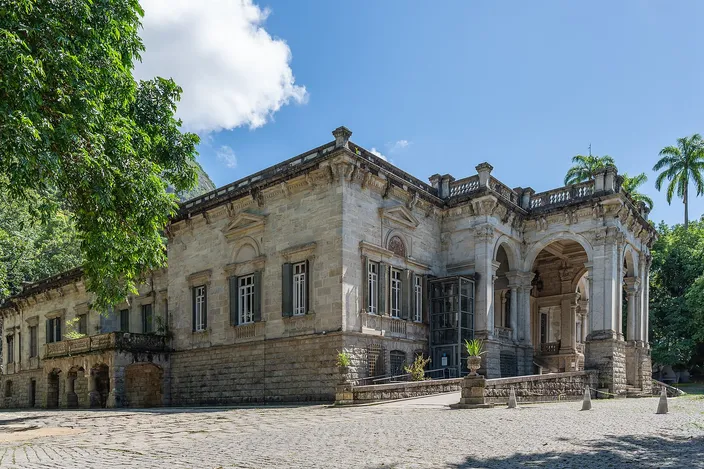 The mansion at the center of Parque Lage on a bright day, a pale two-story villa with an arcaded porch and balustraded roof, seen across an empty cobbled forecourt with dark trees on the left and palms behind it