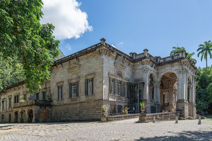 The mansion at the centre of Parque Lage on a bright day, a pale two-storey villa with an arcaded porch and balustraded roof, seen across an empty cobbled forecourt with dark trees on the left and palms behind it