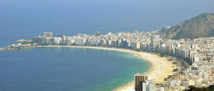 The full crescent of Copacabana seen from a height at the Leme end: pale sand curving away between deep blue water and a solid wall of white apartment blocks, with a forested granite headland in the foreground and green hills closing the far end