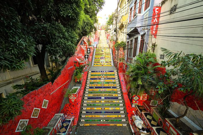 A long staircase covered in yellow, green, blue and red ceramic tiles, seen from the top, with tiled walls and potted plants on both sides