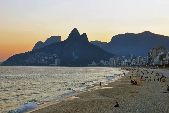 Ipanema beach at sunset seen from the rocks at its eastern end, the sand curving away west towards the twin peaks of a mountain silhouetted against an orange sky, scattered figures on the sand