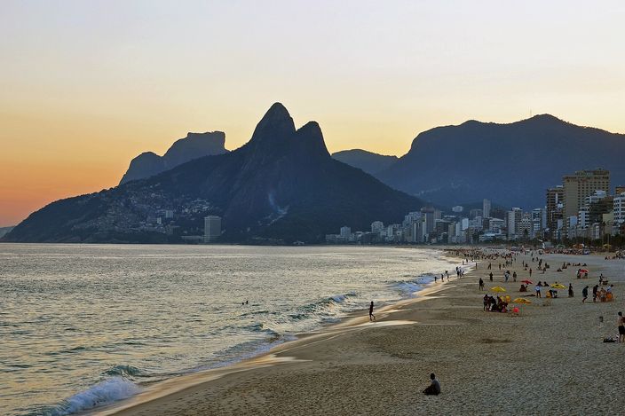 Ipanema beach at sunset seen from the rocks at its eastern end, the sand curving away west towards the twin peaks of a mountain silhouetted against an orange sky, scattered figures on the sand