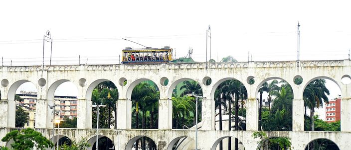 A yellow open-sided tram crossing the top of a two-tier white aqueduct, palm trees behind the arches and people walking on the wet paving below