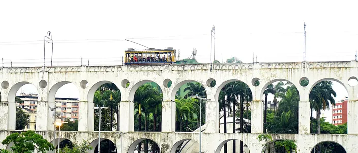 A yellow open-sided tram crossing the top of a two-tier white aqueduct, palm trees behind the arches and people walking on the wet paving below