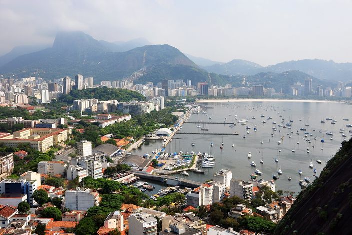 Aerial view of Botafogo: dense low-rise city between forested mountains, curving around a bay full of moored white boats, with Corcovado's hills in the haze behind