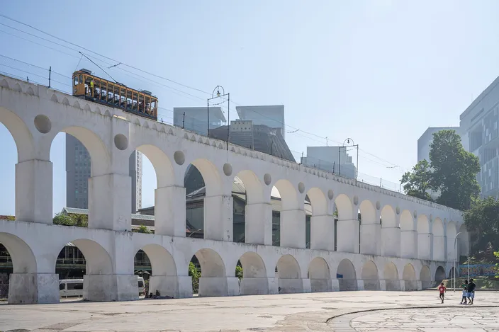 The yellow bondinho tram crossing the tall white Arcos da Lapa aqueduct arches, with modern towers of Centro behind under a clear sky