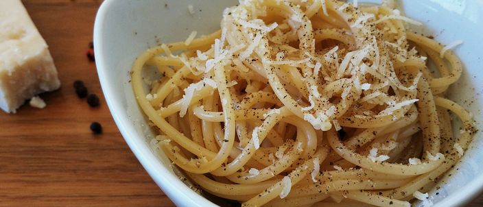 A white bowl of spaghetti cacio e pepe topped with grated pecorino and cracked black pepper, beside a wedge of hard cheese and scattered peppercorns