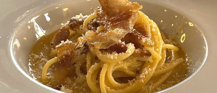 A nest of spaghetti alla carbonara in a white bowl, the sauce pale gold and glossy, topped with shards of crisp guanciale and a dusting of grated pecorino.