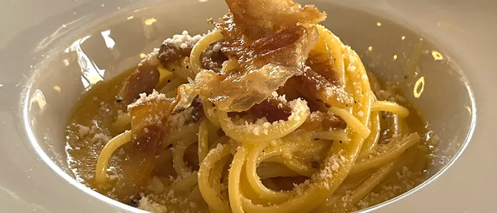 A nest of spaghetti alla carbonara in a white bowl, the sauce pale gold and glossy, topped with shards of crisp guanciale and a dusting of grated pecorino.