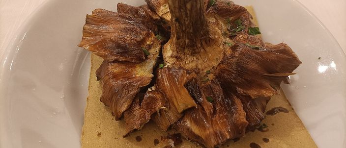 A whole deep-fried artichoke pressed open into a flower shape, its outer leaves splayed golden and crisp, served on a plate lined with kraft paper