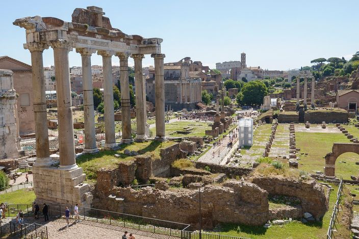 The Roman Forum seen from above at its western end: eight granite columns of the Temple of Saturn standing under an inscribed entablature in the left foreground, the excavated field of brick and stone foundations running away east, a row of column stumps marking the length of a basilica along the right, umbrella pines and a square medieval brick tower on the ridge behind, and small groups of visitors on the gravel paths.