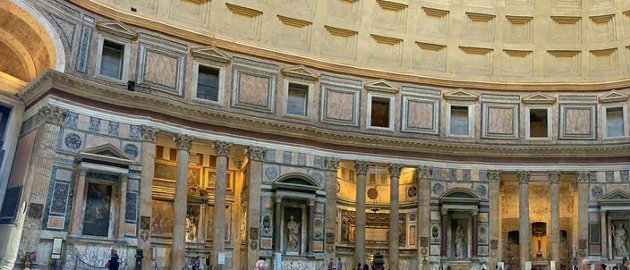 The interior of the Pantheon in warm daylight: a wide curved band of the marble-clad drum with columned aediculae and their triangular and segmental pediments, dark niches holding framed altarpieces between them, and the lowest rows of the coffered concrete dome springing above the entablature.