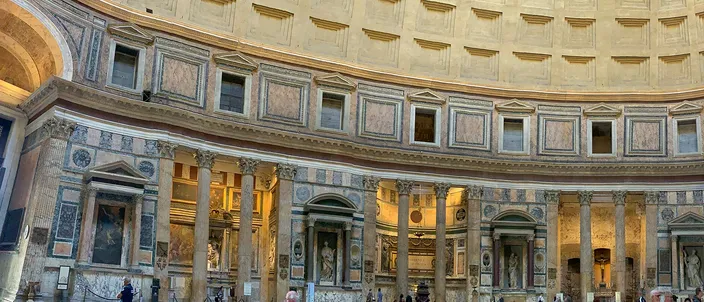 The interior of the Pantheon in warm daylight: a wide curved band of the marble-clad drum with columned aediculae and their triangular and segmental pediments, dark niches holding framed altarpieces between them, and the lowest rows of the coffered concrete dome springing above the entablature.