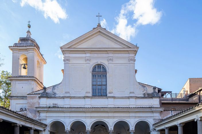 The white eighteenth-century facade of the Basilica of San Clemente rising behind its arcaded courtyard, an open belfry on a small tower to the left, a stone fountain standing on the cobbled atrium floor and potted cycads under the portico arches, in low sun against a blue sky.