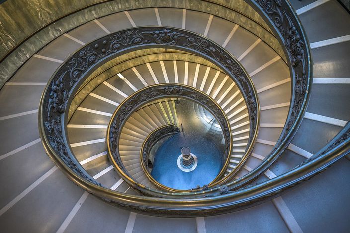 The double-helix staircase of the Vatican Museums photographed straight down its well: two intertwined ramps of pale steps spiralling inward around an ornate bronze balustrade, narrowing turn by turn to a single lit lamp at the bottom.