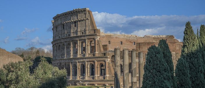 The Colosseum in low afternoon sun seen across the edge of the archaeological park, its tiers of arches in warm travertine above a slope of olive trees, with three standing columns of a ruined temple and a line of dark cypresses in front of it under a blue sky with broken cloud.