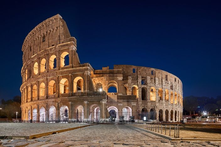 The Colosseum at blue hour, empty of people, its travertine arcades lit warm gold against a deep blue sky, with the paved forecourt running to the base of the ruined outer wall.