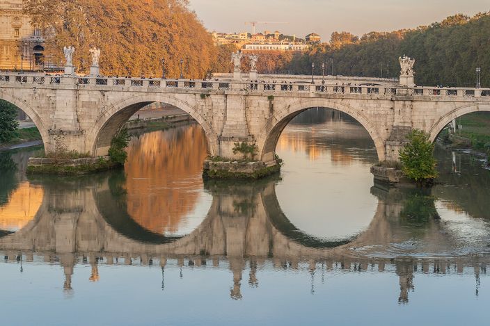 Ponte Sant'Angelo in low golden light, its stone arches and their reflections forming full circles on the still Tiber, Bernini's angel statues ranged along both parapets, autumn foliage on the far bank.
