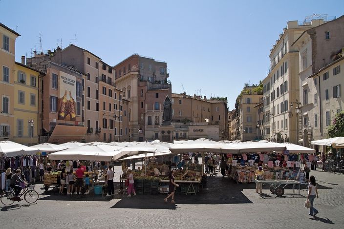The Campo de' Fiori market in full sun: rows of white canvas awnings over produce and clothing stalls filling the square, ochre and cream palazzo facades on all sides, and the dark hooded bronze of Giordano Bruno standing on its plinth in the middle.