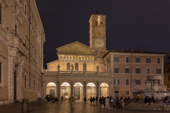 The facade of Santa Maria in Trastevere floodlit at night, its gold-ground mosaic frieze of figures glowing above the portico, the romanesque bell tower with its clock rising behind, and people sitting on the fountain steps in the square below.
