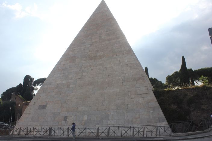 The Pyramid of Cestius: a steep white marble-faced pyramid rising straight from the pavement behind iron railings, cypresses and umbrella pines on the bank beside it, a single passer-by at its foot for scale.