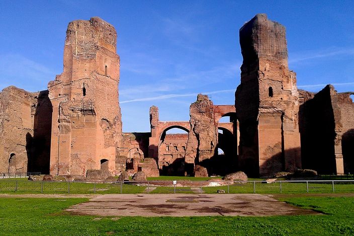 The Baths of Caracalla in low sun: two towering fingers of bare red brickwork standing thirty metres high with arched openings between them, deep blue sky behind, mown grass and the flat footprint of the vanished halls in front.