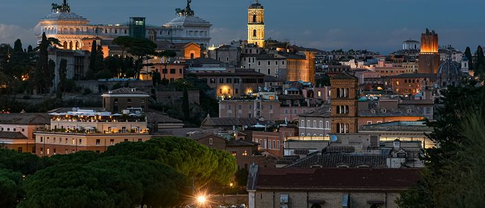 Rome's rooftops at blue hour, seen from a hill: terracotta roofs and umbrella pines in the foreground, the floodlit white bulk of the Vittoriano with its bronze quadrigae on the skyline, and the bell tower of the Palazzo Senatorio beside it.