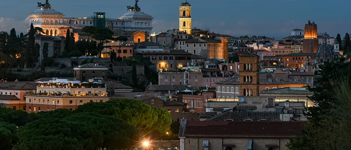 Rome's rooftops at blue hour, seen from a hill: terracotta roofs and umbrella pines in the foreground, the floodlit white bulk of the Vittoriano with its bronze quadrigae on the skyline, and the bell tower of the Palazzo Senatorio beside it.