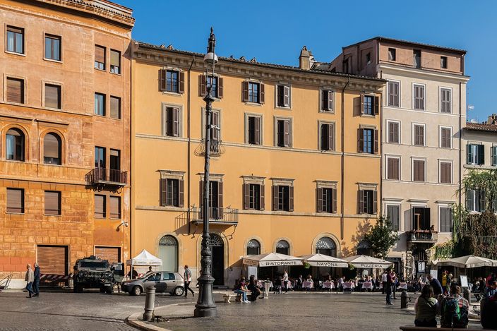 A sunlit ochre and terracotta palazzo facade on Piazza Navona, with an ornate lamp-post, shuttered windows, cafe awnings and people sitting on the paving in the foreground