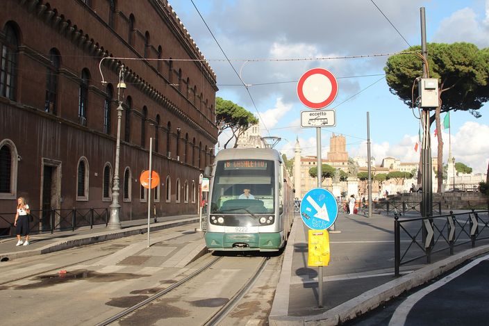 A green-and-white ATAC tram stopped on the tracks beside the long brick facade of Palazzo Venezia, with umbrella pines, an Italian flag and a no-entry sign, the ruins of Trajan's Forum in the distance.