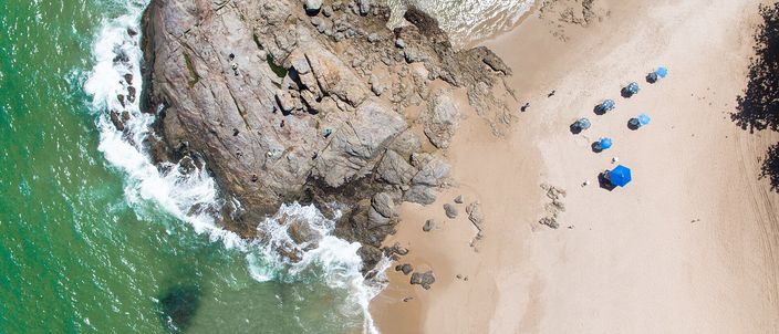 Aerial view of a dark rocky point where waves break white against it, meeting a curve of pale sand with a small cluster of blue beach umbrellas, Itapuã beach, Salvador.