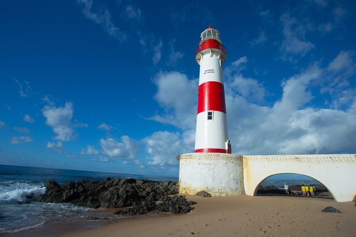 The red-and-white banded Ponta de Itapuã lighthouse standing on a rocky outcrop above the sand, beside a whitewashed archway walkway, with a few people at its base.