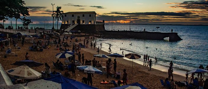 Forte de Santa Maria silhouetted against an orange sunset sky, with a crowd still gathered on the sand of Porto da Barra as the sun sets into the calm bay.