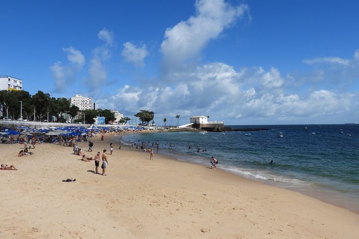 Beachgoers on the calm, gently sloping sand of Porto da Barra in daylight, turquoise water lapping the shore with the white walls of Forte de Santa Maria visible at the far point.