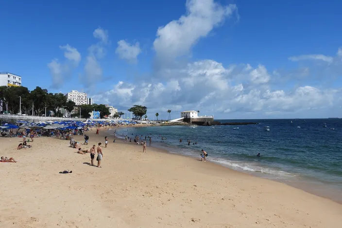 Beachgoers on the calm, gently sloping sand of Porto da Barra in daylight, turquoise water lapping the shore with the white walls of Forte de Santa Maria visible at the far point.