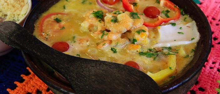 A black clay pot of orange moqueca baiana with prawns, rings of red pepper and coriander, on a colourful crocheted mat.