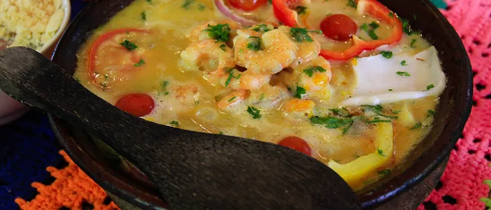 A black clay pot of orange moqueca baiana with prawns, rings of red pepper and cilantro, on a colorful crocheted mat.