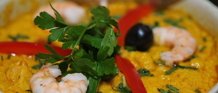 A white bowl of smooth orange-gold vatapá garnished with whole prawns, parsley, strips of red pepper and a black olive.