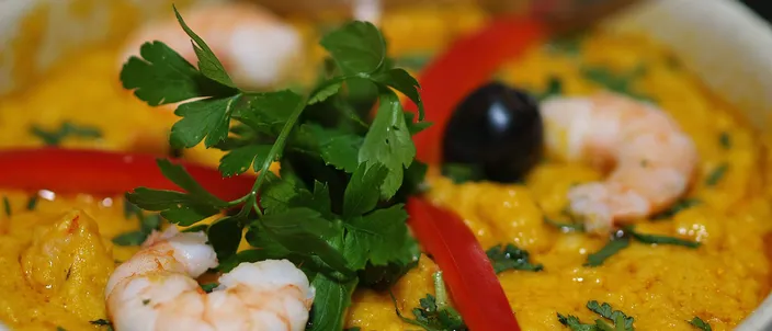 A white bowl of smooth orange-gold vatapá garnished with whole prawns, parsley, strips of red pepper and a black olive.