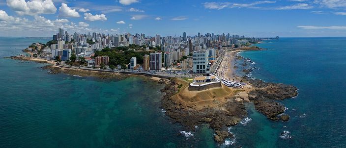 Aerial panorama of the Barra peninsula tip in Salvador, the Farol da Barra fort and lighthouse jutting into turquoise Atlantic water with the city skyline curving behind under a blue sky