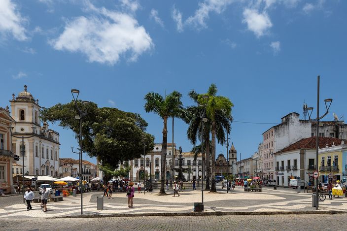 Terreiro de Jesus in Salvador on a bright day: a large square paved in black-and-white wave mosaic, tall palms and a stone fountain in the middle, white and pastel church façades and colonial houses closing the square on three sides, people crossing in scattered groups