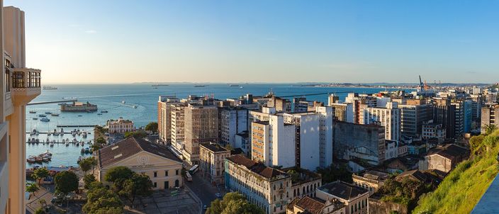 View from a high parapet over Salvador's lower city at golden hour: colonial rooftops and white apartment blocks stepping down to a quay with moored boats, the wide blue Baia de Todos os Santos beyond with ships at anchor and port cranes on the far right
