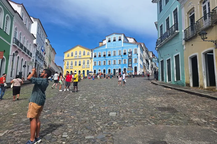 Largo do Pelourinho in Salvador seen from its top on a bright day: a broad cobbled square sloping downhill between rows of pastel colonial houses, the tall blue facade of the Fundacao Casa de Jorge Amado closing the view at the far end beside a yellow one, visitors scattered across the stones